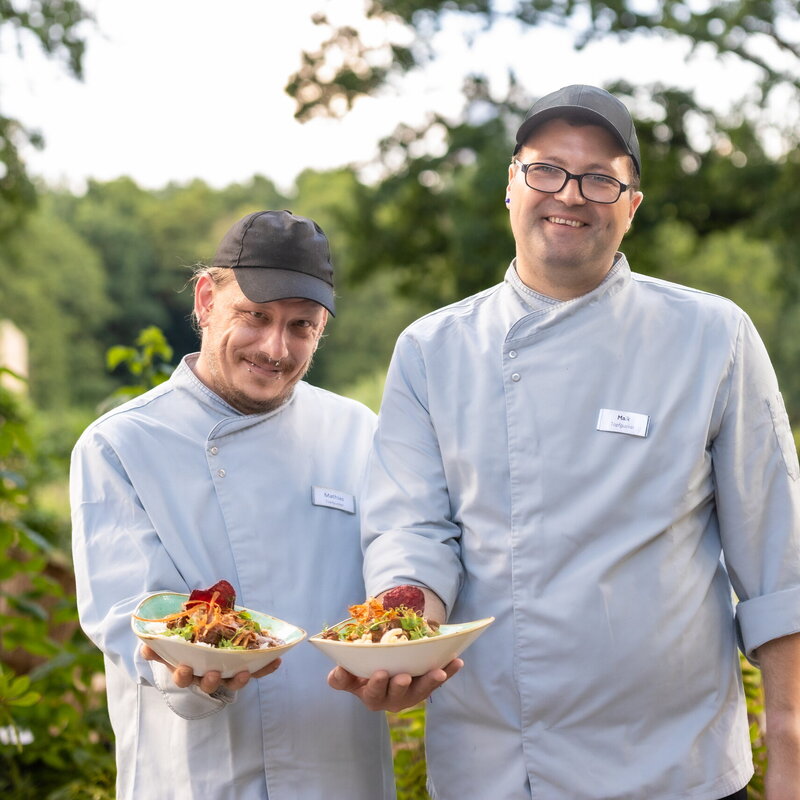 Zwei Mitarbeiter des Bistros halten Schüsseln mit Gerichten in die Kamera